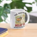 White ceramic mug printed with cartoon cockatoo eating tomato-sauced meat pie under gum-tree backdrop